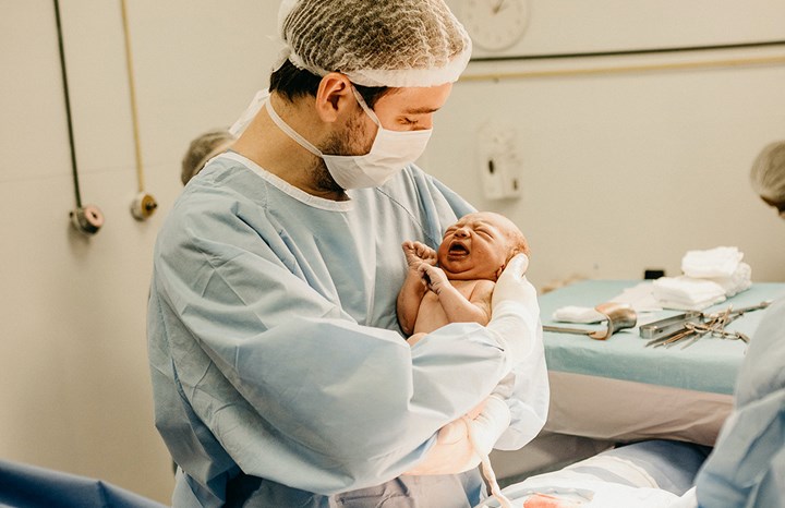 Pappa håller sitt nyfödda barn på BB. Han bär sjukhuskläder och de är i förlossningsrummet.