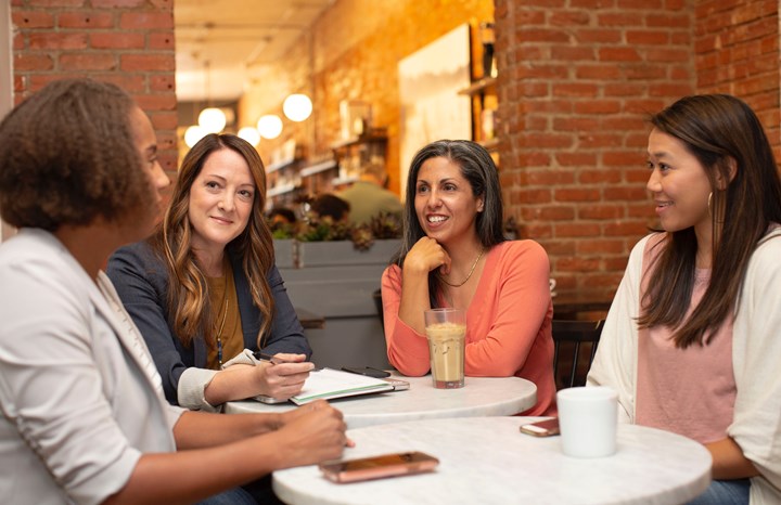 Fyra kvinnor sitter runt ett bord på ett café, med telefoner, kaffekoppar och anteckningsblock framför sig. 