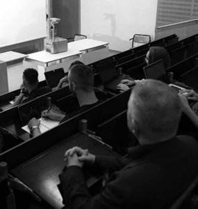 En sluttande föreläsningssal i universitetsmiljö där studenter sitter i åhörarstolar och en kvinna står längst fram och talar. 